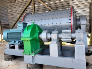 Industrial machine with gray cylindrical drum and visible components in blue and green, set on a metal frame against a corrugated background.