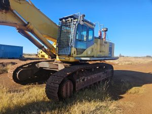 A yellow Komatsu hydraulic excavator with a protective cage over the cab sits idle on a dirt site with grass in the foreground.