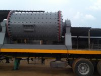 A large, industrial gray ball mill with a studded surface is securely mounted on a yellow flatbed trailer, conveying a sense of robust engineering.